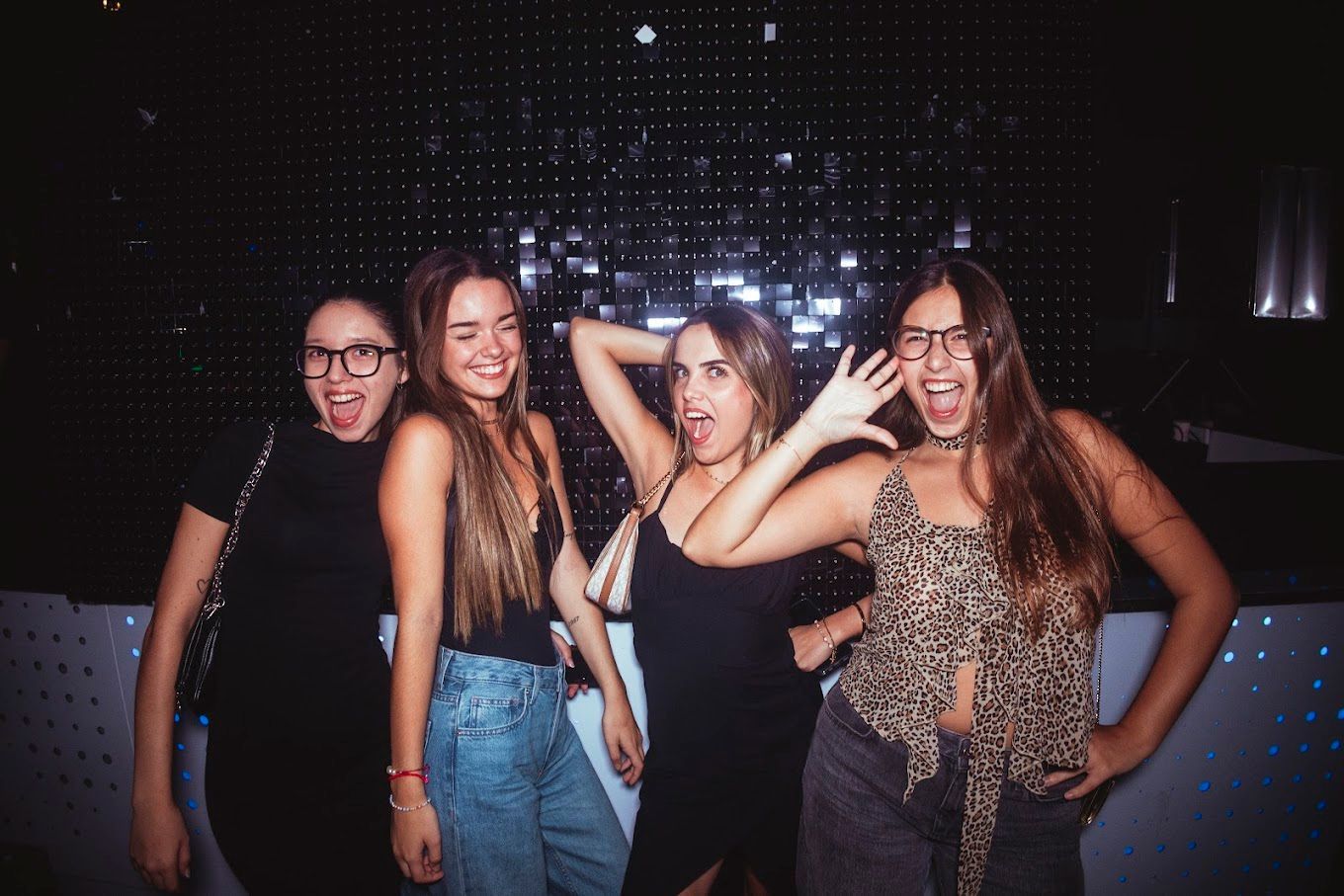 Cuatro jóvenes sonríen y posan en una discoteca. Llevan tops negros con estampado de leopardo. Fondo oscuro con luces.