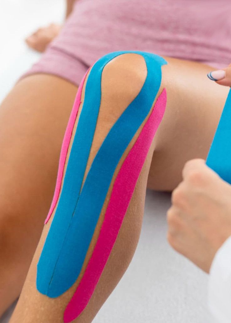 Rodilla con cinta kinesiológica azul y rosa aplicada por una persona con bata blanca.