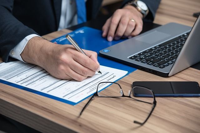 Un hombre está firmando un documento frente a una computadora portátil.