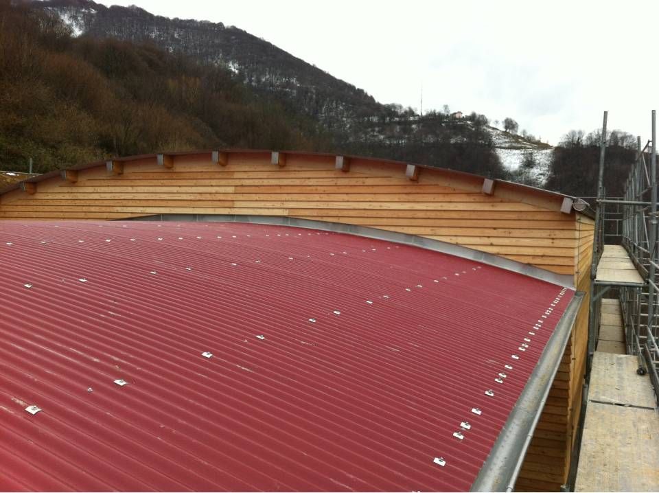 Tetto in lamiera ondulata rossa su un edificio curvo in legno con una collina innevata sullo sfondo.