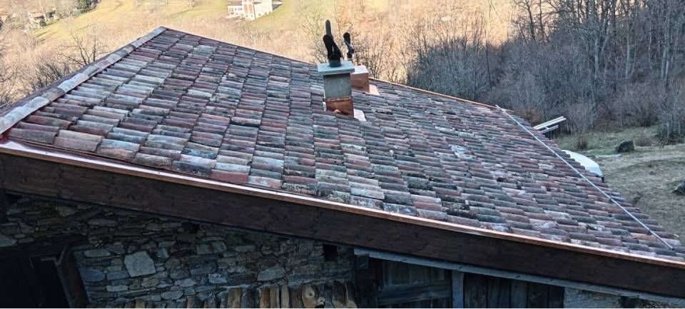 Un rustico edificio in pietra con un tetto di tegole rosse e un camino in un paesaggio montuoso e boscoso.
