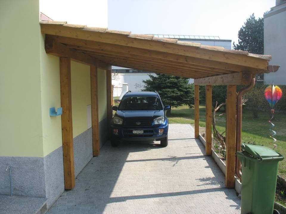 Carport in legno con un'auto blu parcheggiata sotto. Cestino verde e decorazioni colorate.