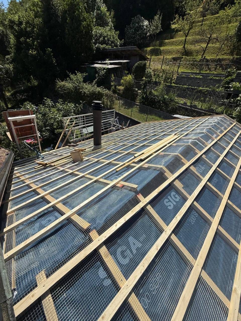 Tetto in costruzione, con membrana nera, travi in legno e camino. Sullo sfondo alberi e pendio.