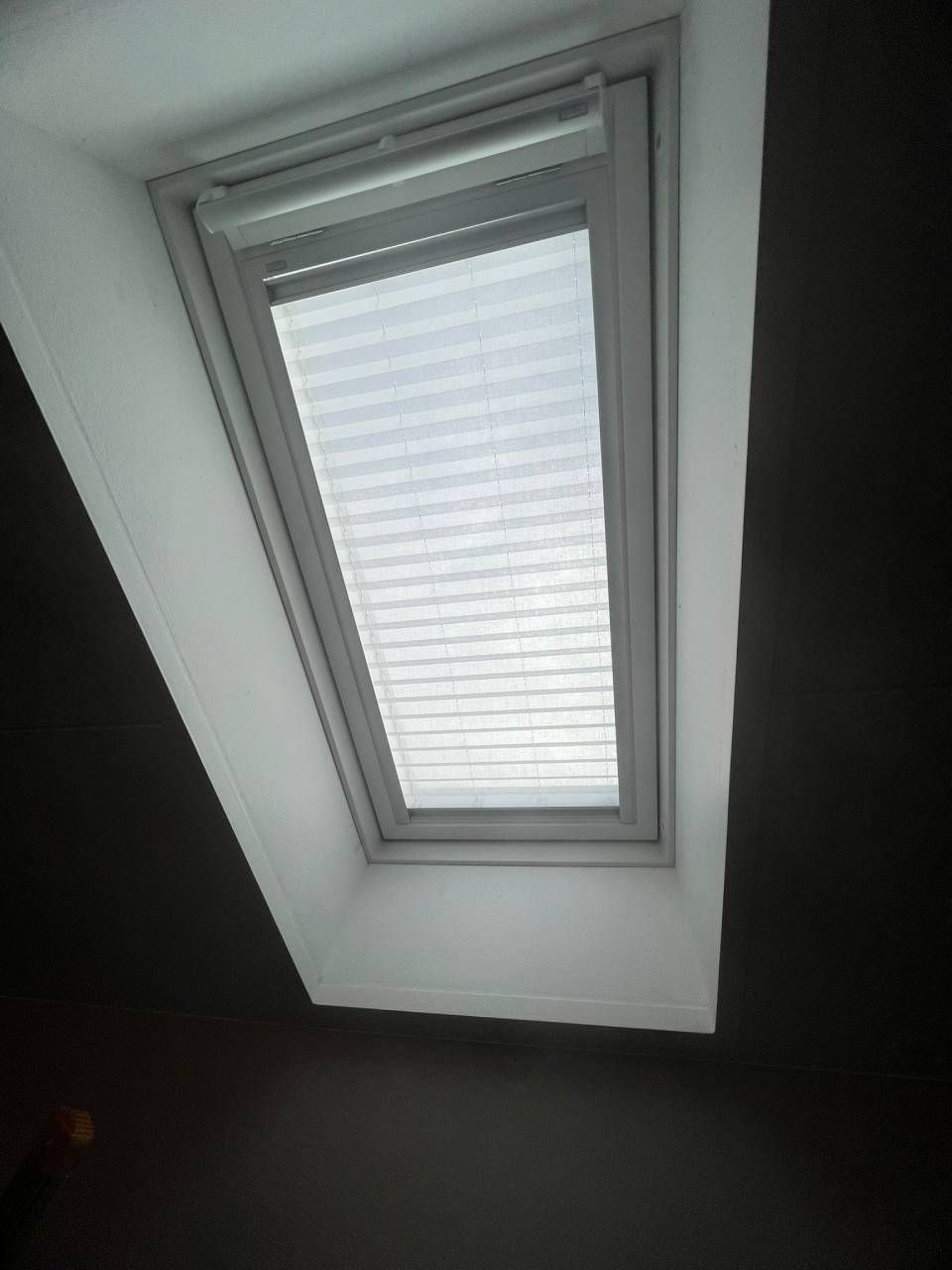 Lucernario con tenda plissettata bianca in una stanza dalle pareti scure.