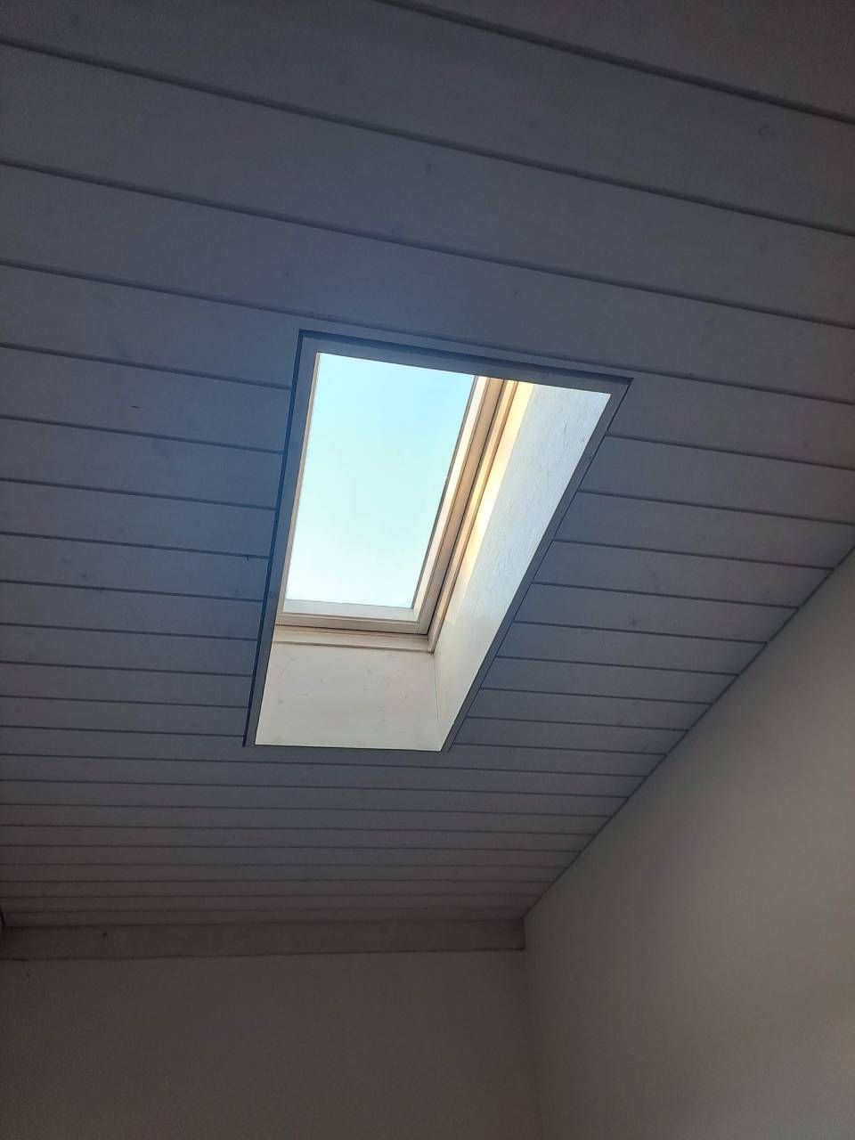 Lucernario in un soffitto spiovente rivestito di pannelli grigi, che incornicia la vista del cielo azzurro.