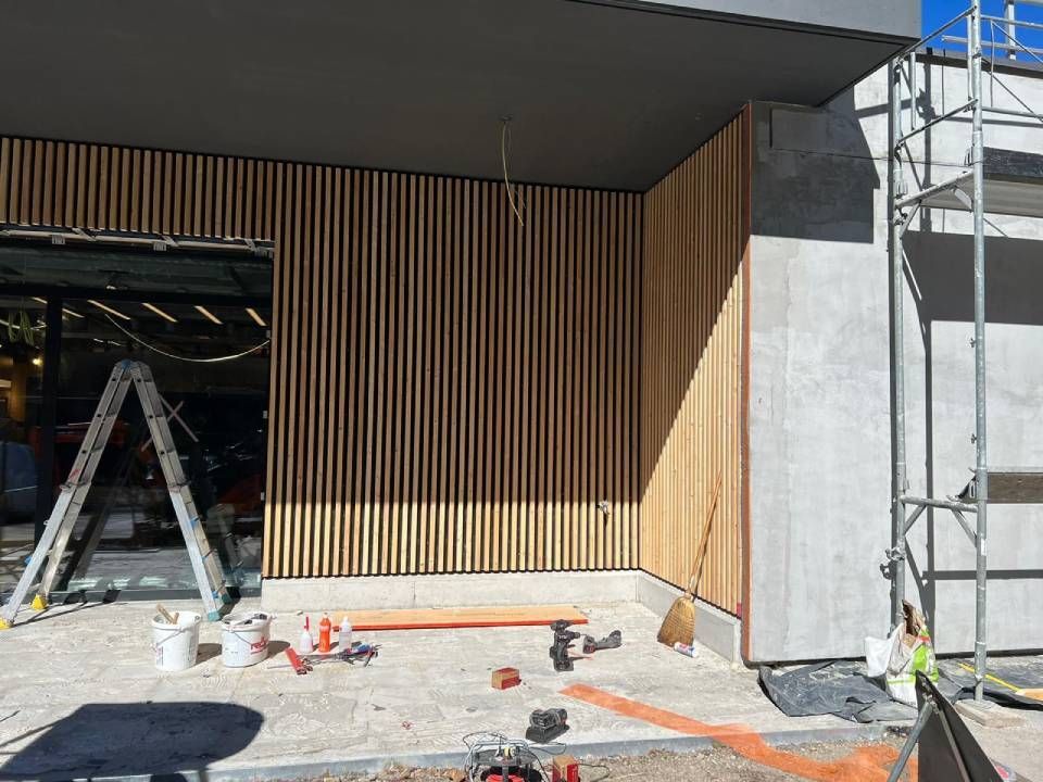 Pannelli in doghe di legno sulla facciata esterna di un edificio in costruzione; sono visibili impalcature, attrezzi e secchi di vernice.