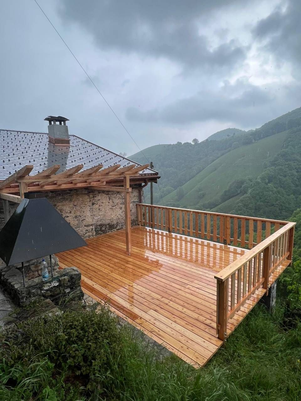Ponte di legno attaccato a un edificio in pietra, con vista su una catena montuosa verdeggiante sotto un cielo nuvoloso.