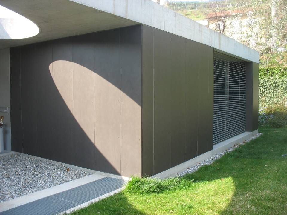 Edificio moderno con pannelli marroni, tetto in cemento e una finestra con grate. La luce del sole proietta una grande ombra.