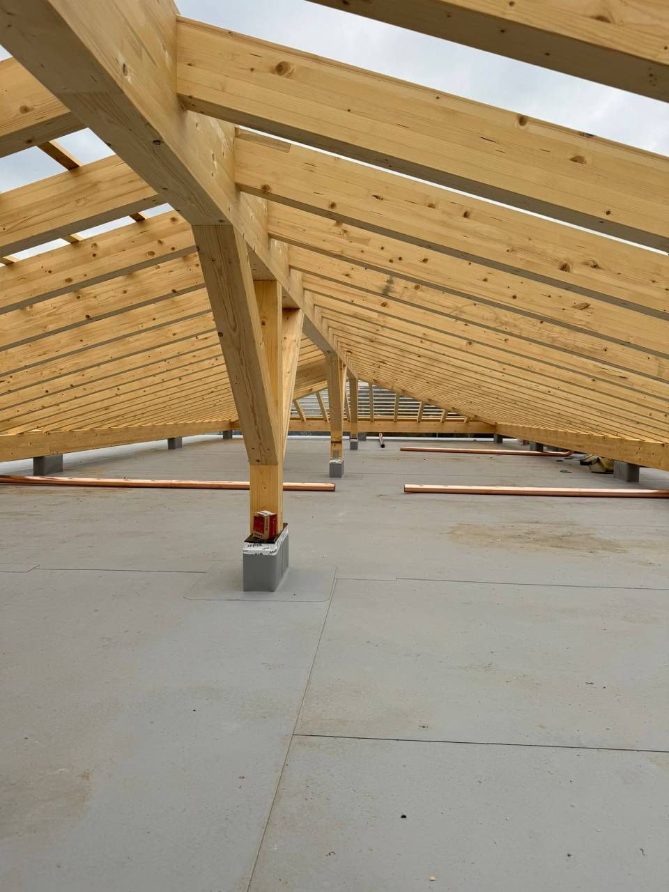 Vista interna dell'edificio incompiuto con travi in legno, pavimento in cemento e pali di sostegno.