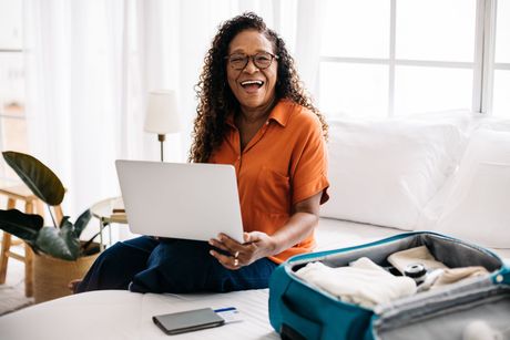 Une dame souriante devant son ordinateur, son téléphone et une valise ouverte