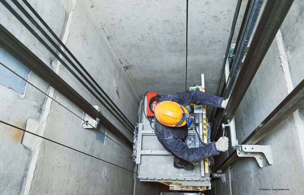 Arbeiter mit orangefarbenem Schutzhelm bedient einen Aufzug in einem engen Betonaufzugsschacht