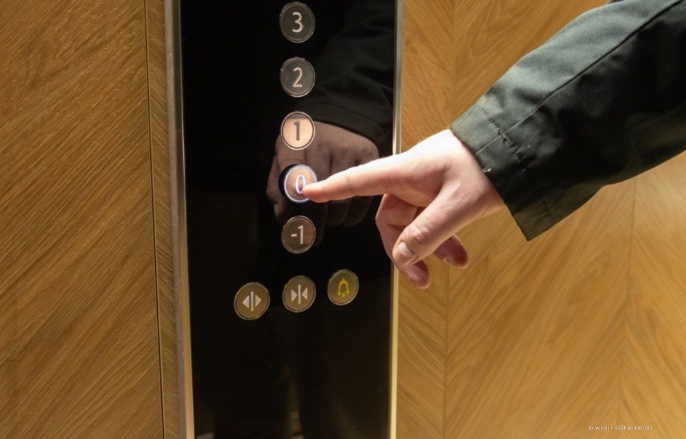 Eine Hand drückt ein Aufzugsknopffeld mit beleuchteten Stockwerksnummern und goldenen Bedienknöpfen.