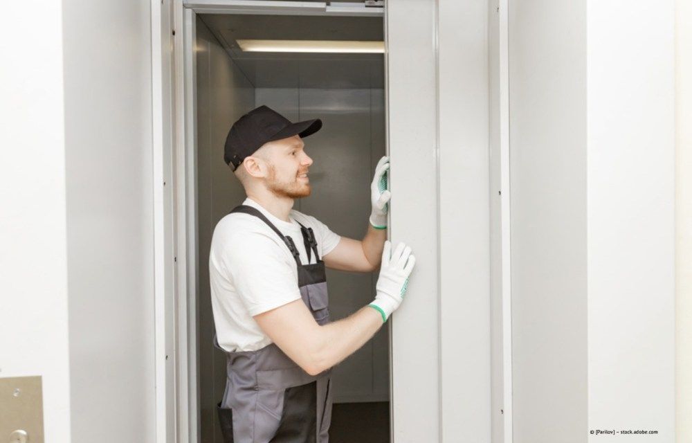 Ein Arbeiter mit schwarzer Kappe und Schürze misst in einem hellen Raum mit einem Maßband eine Türöffnung aus.