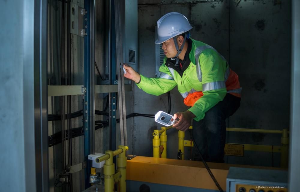 Ein Arbeiter mit Schutzhelm und Sicherheitsweste prüft elektrische Anlagen in einem schwach beleuchteten Industrieraum.