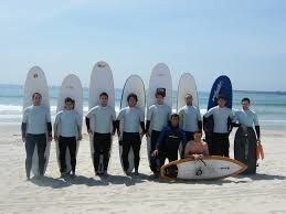 Group of surfers posing with surfboards on a sunny beach, one sitting on a board in front.