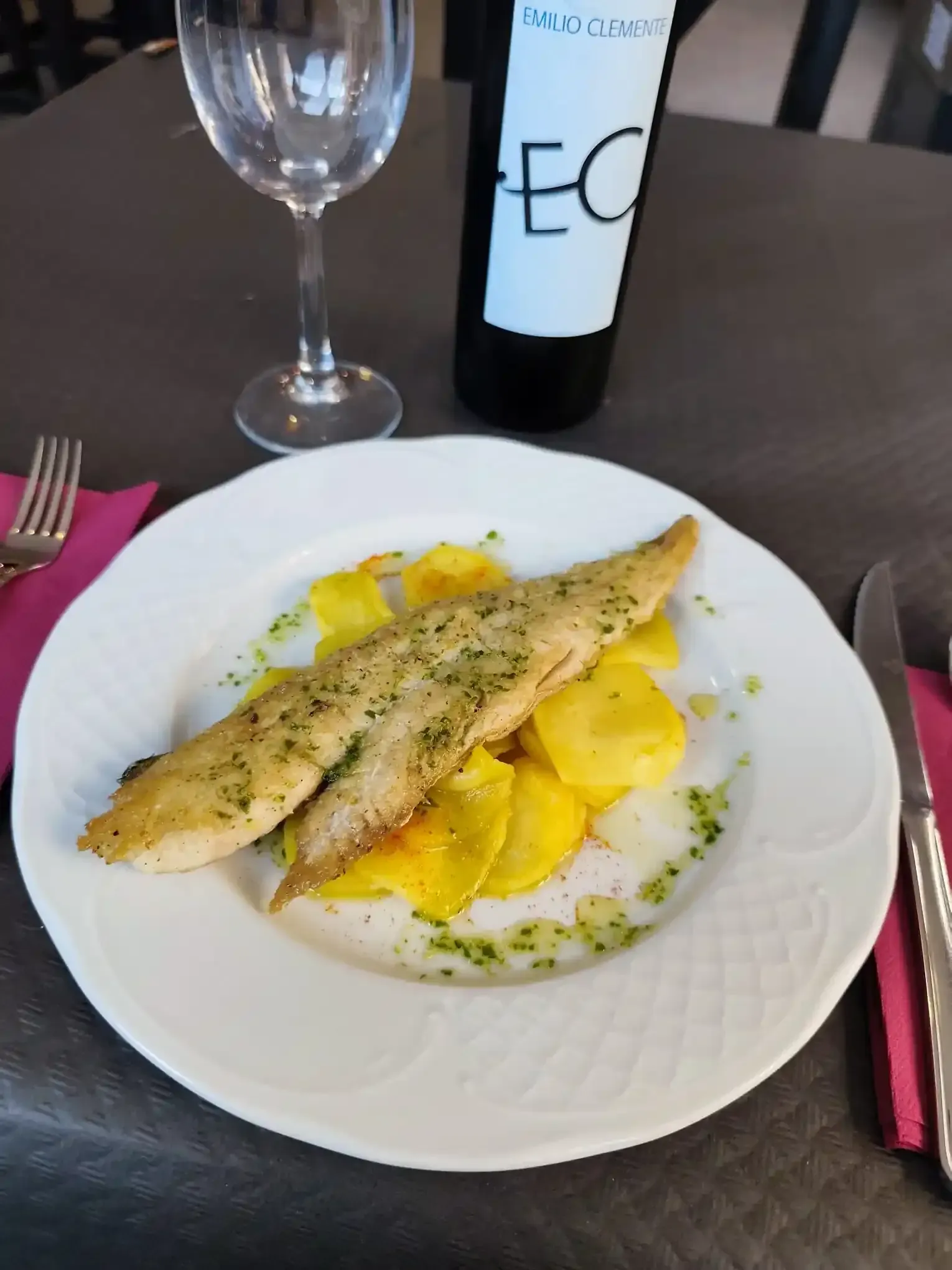 Filetes de pescado con patatas en un plato blanco, copa de vino, botella, tenedor y cuchillo sobre una mesa.