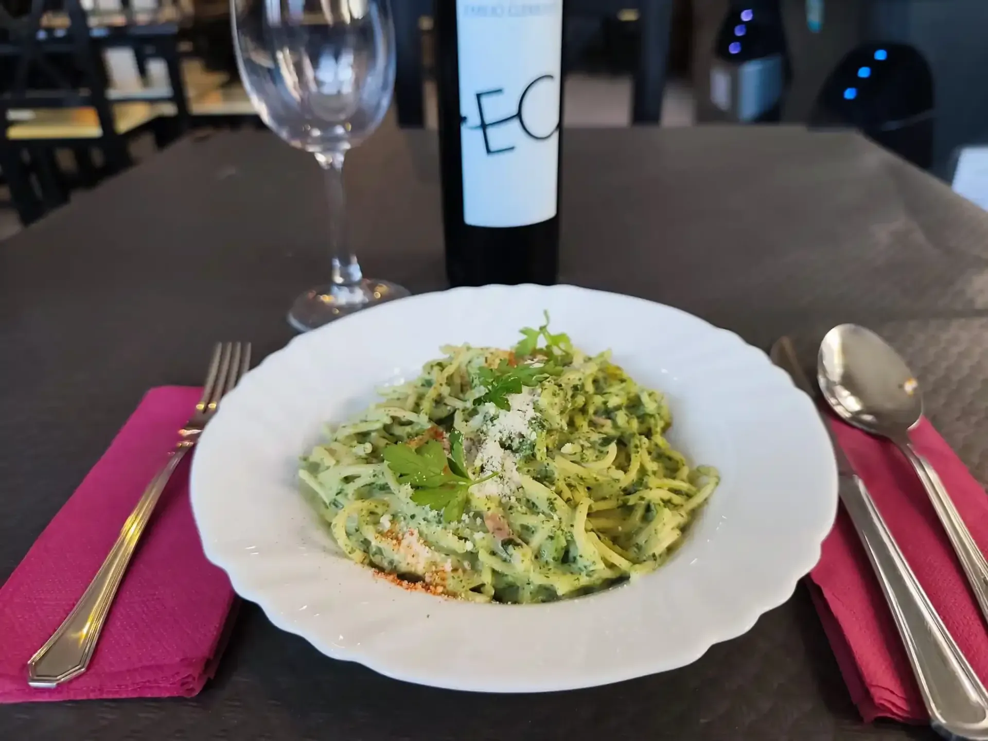 Plato de pasta en un plato blanco con copa de vino, cubiertos y una botella de vino sobre una mesa con una servilleta.