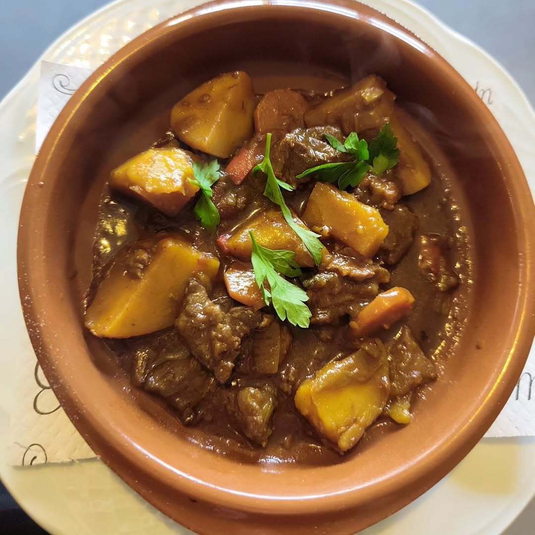 Guiso de carne de res en recipiente marrón, con guarnición de patatas, zanahorias y perejil.