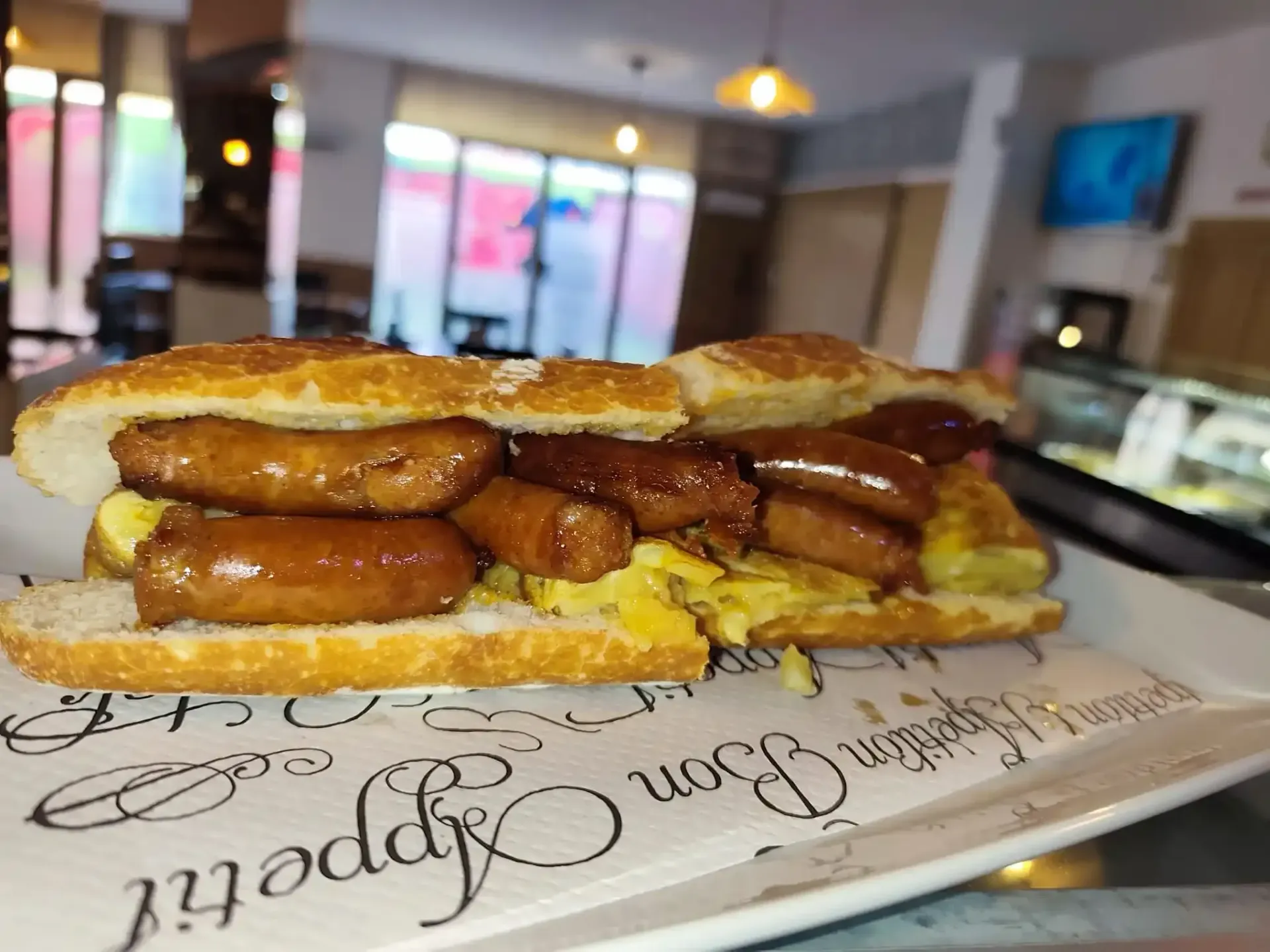 Sándwich de salchicha en un plato blanco con texto, en un entorno de cafetería.