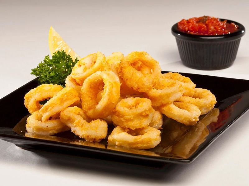 Anillos de calamares fritos en un plato negro con salsa roja, rodaja de limón y perejil.