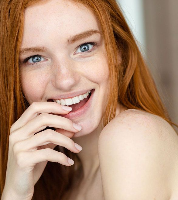 Una mujer de cabello rojo y ojos azules sonríe con la mano en la boca.