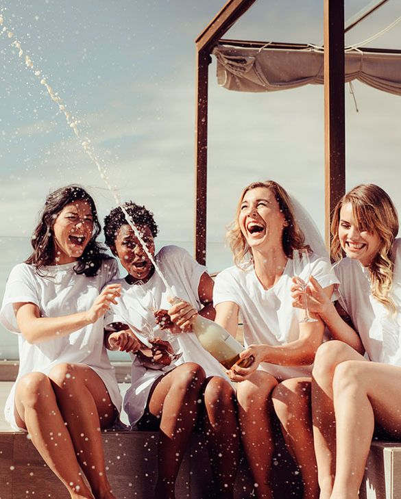 Un grupo de mujeres están sentadas en un banco sosteniendo botellas de champán y riendo.