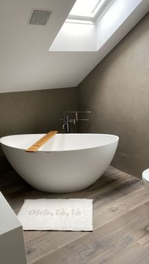 Eine weisse, rechteckige freistehende Badewanne mit modernem Chromhahn, die auf einem dunklen Holzboden in einem Raum mit schrägen Wänden steht.