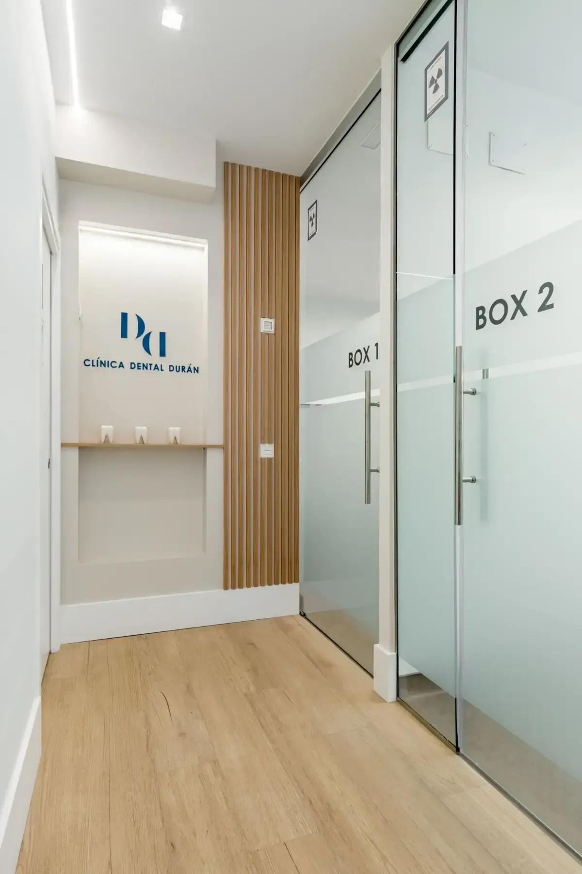 Pasillo de una clínica médica con dos salas de examen con puertas de vidrio, piso de madera y el logotipo de la clínica.