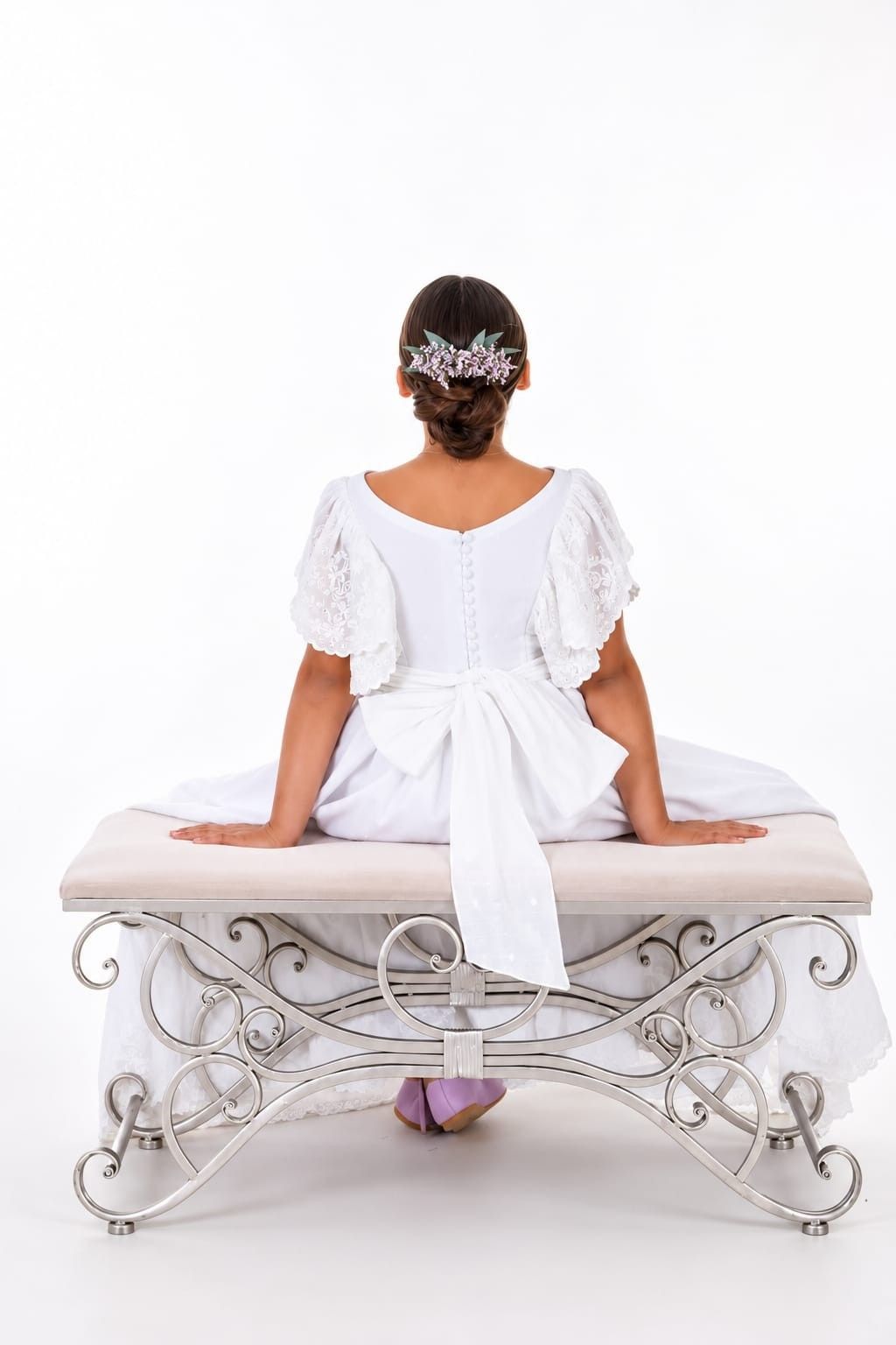 Mujer con vestido blanco sentada en un banco ornamentado, vista desde atrás, sobre un fondo blanco.