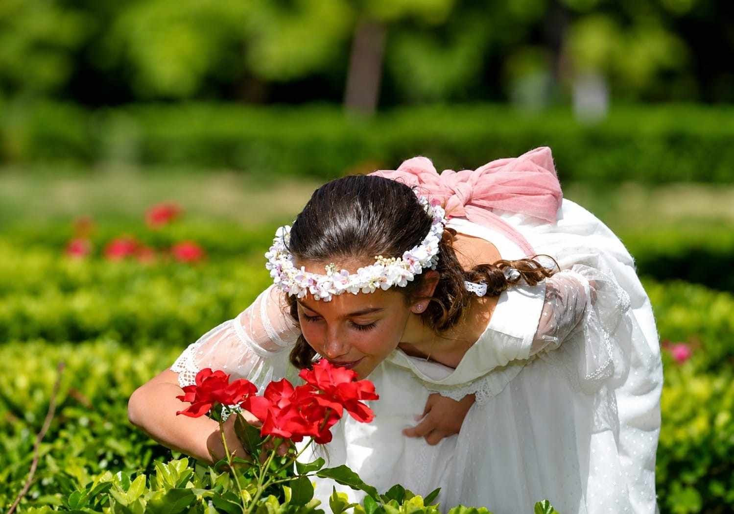 Una joven con un vestido blanco huele flores rojas en un jardín verde.