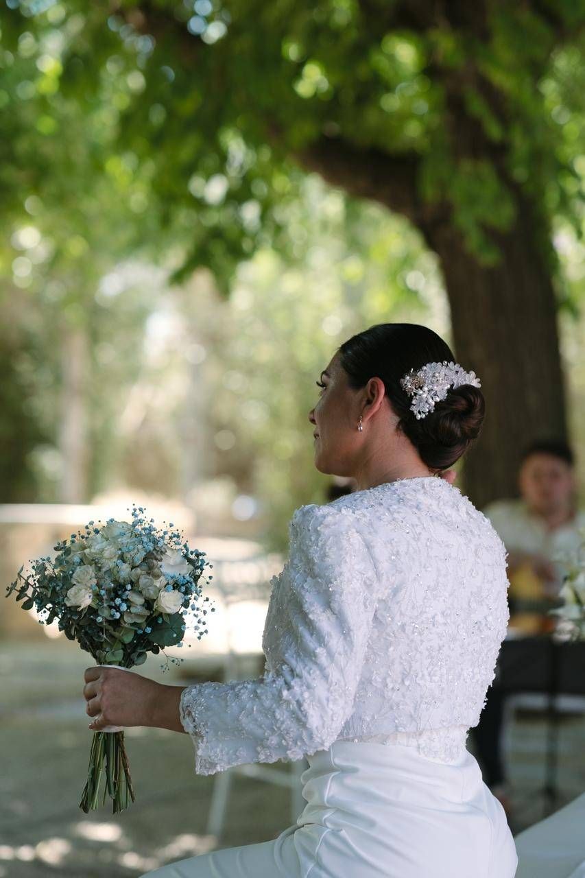 Novia con vestido blanco sosteniendo un ramo al aire libre bajo los árboles.