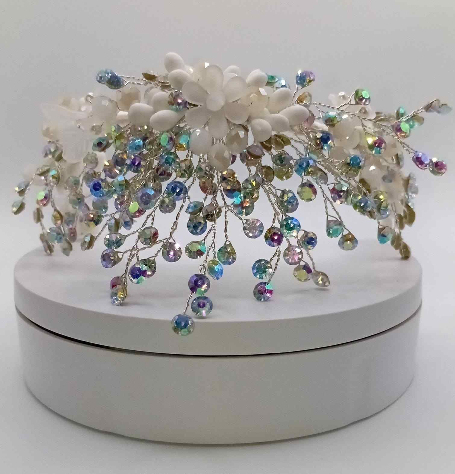 Accesorio para el cabello blanco con flores y joyas, con cuentas iridiscentes, en una caja redonda blanca.