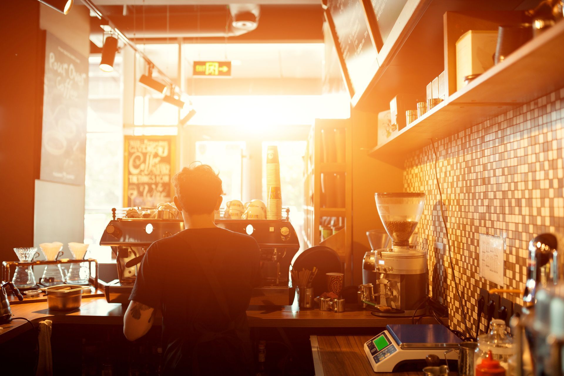 Un barista derrière sa machine à café, baigné par la lumière vive du soleil dans un café.