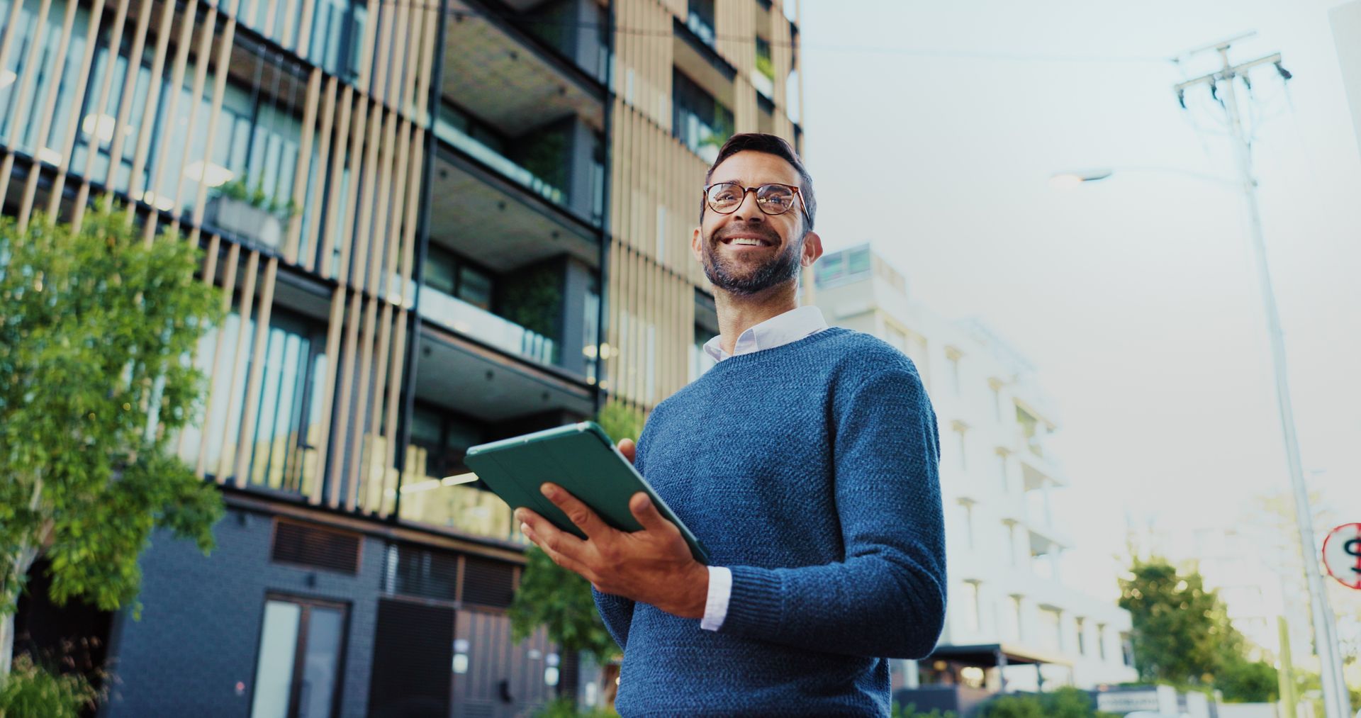 Un homme portant des lunettes tient une tablette et sourit devant un immeuble d'appartements moderne.