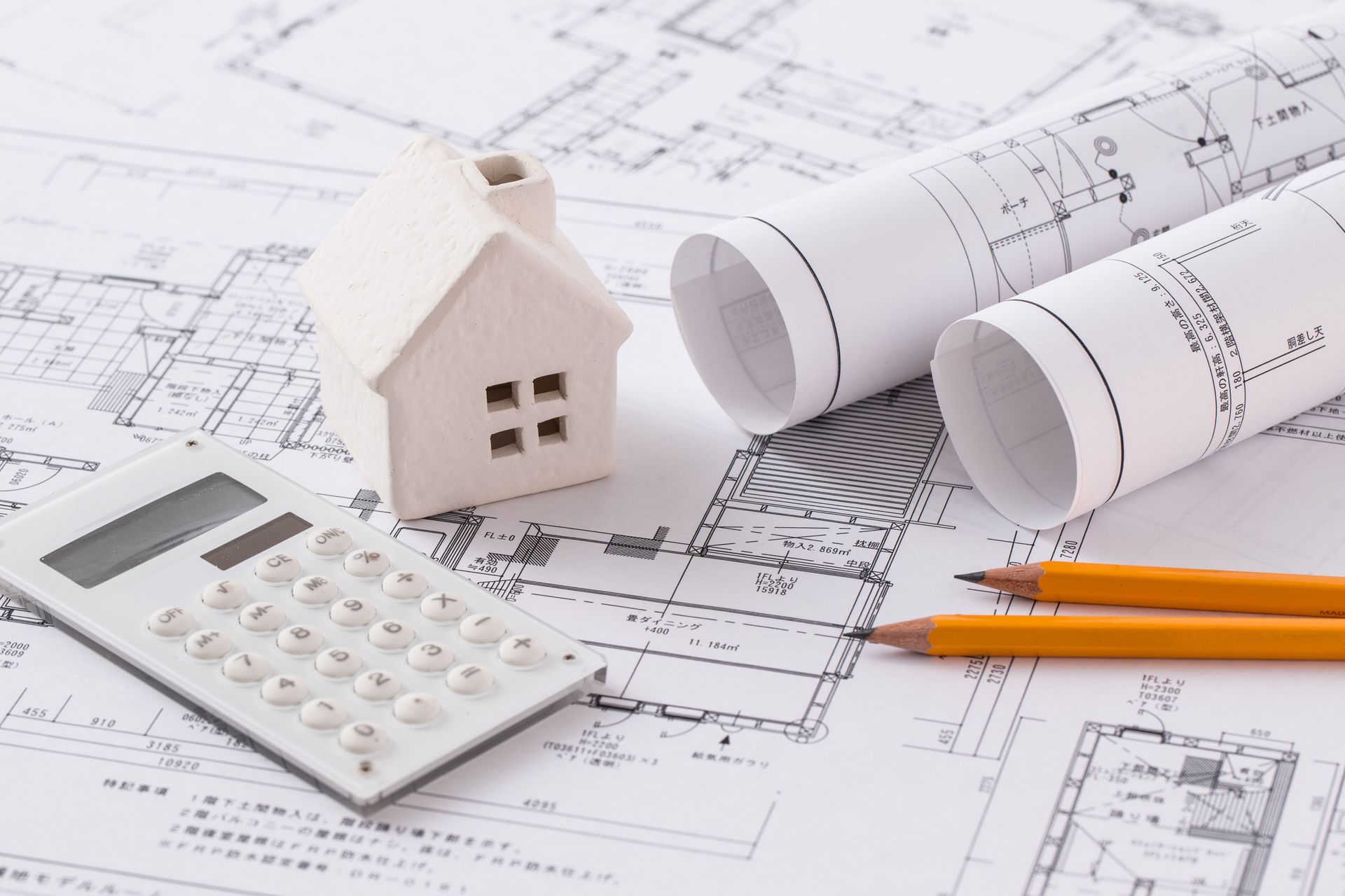 Maison miniature, plans, calculatrice et crayons sur un bureau, représentant la planification de la construction et les calculs financiers.