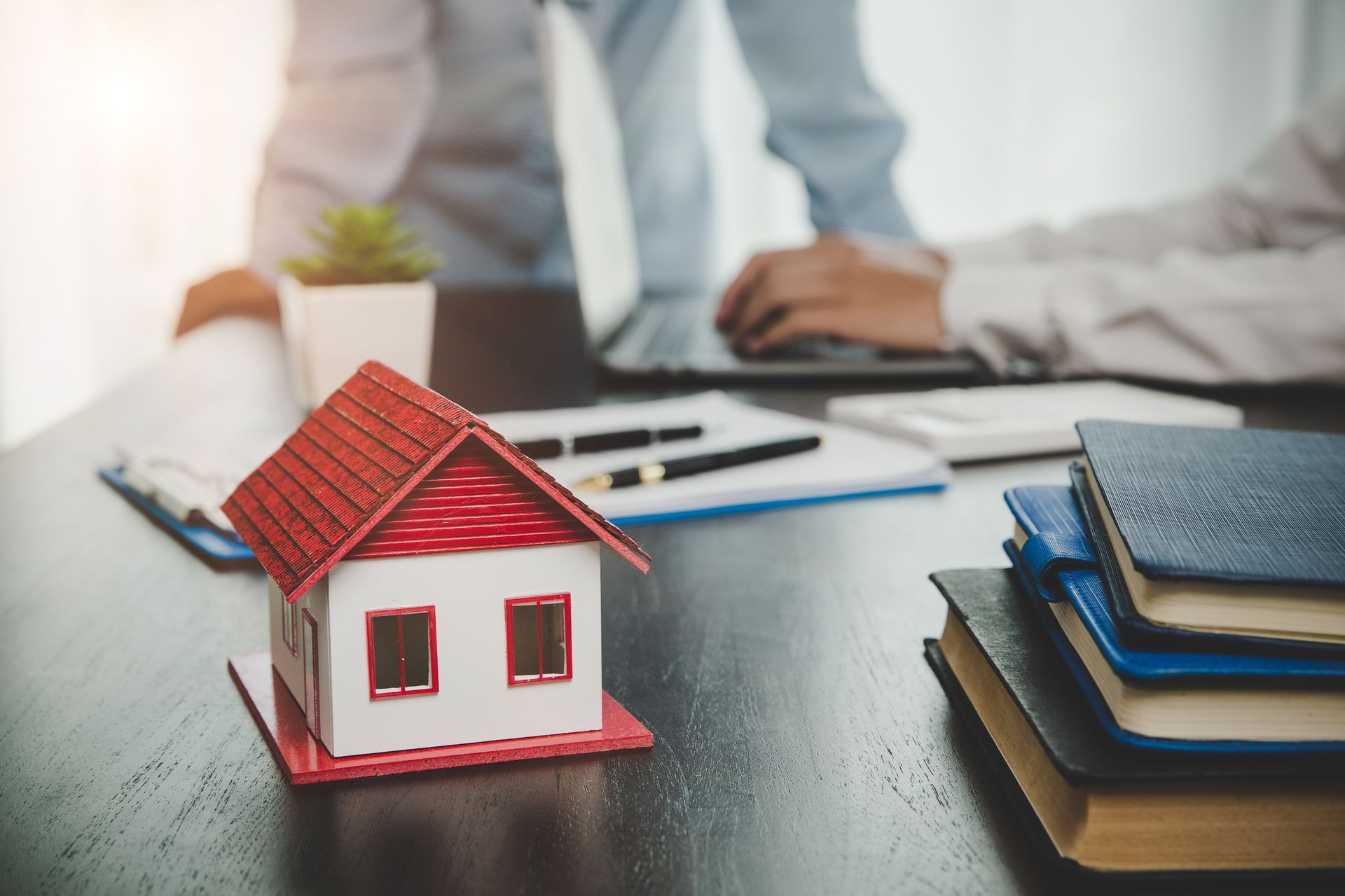 Une maison miniature posée sur une table, avec une personne utilisant un ordinateur portable et des documents, probablement en pleine discussion immobilière.