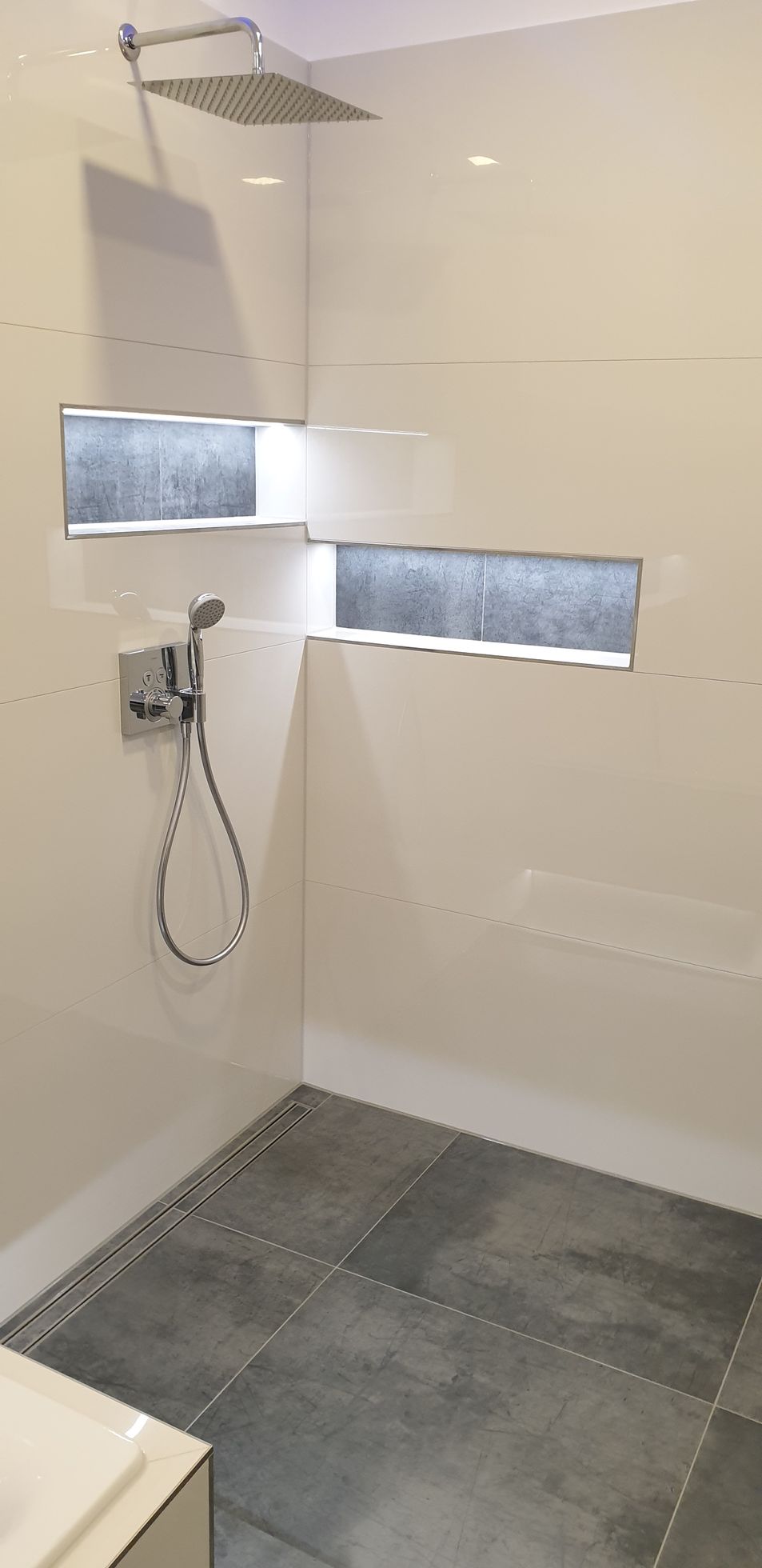 Eine moderne, geflieste Dusche mit zwei beleuchteten Ablagen. Der Boden ist grau gefliest.