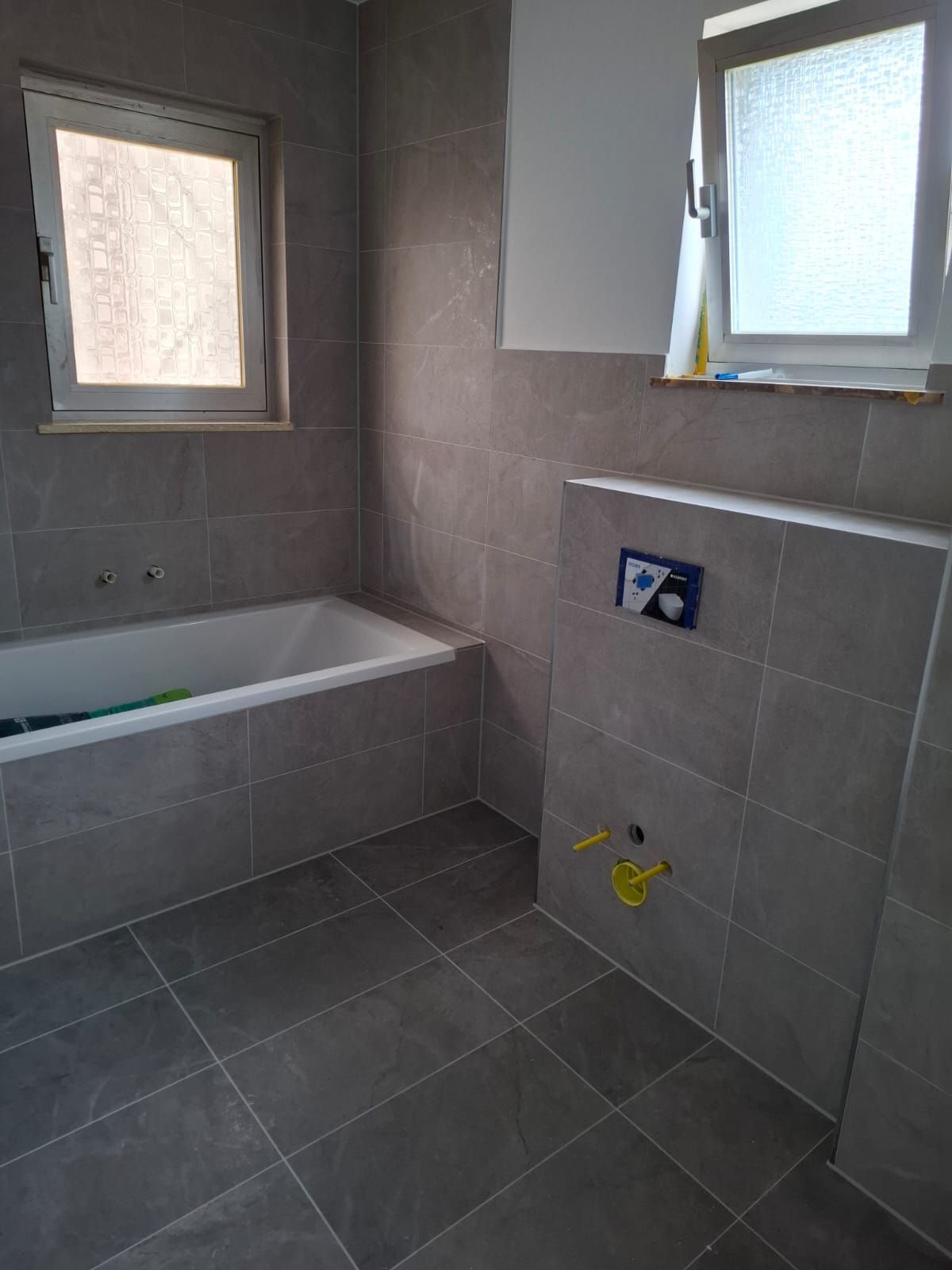 Grau gefliestes Badezimmer mit Badewanne, zwei Fenstern und Toilette an einer halbhohen Wand.