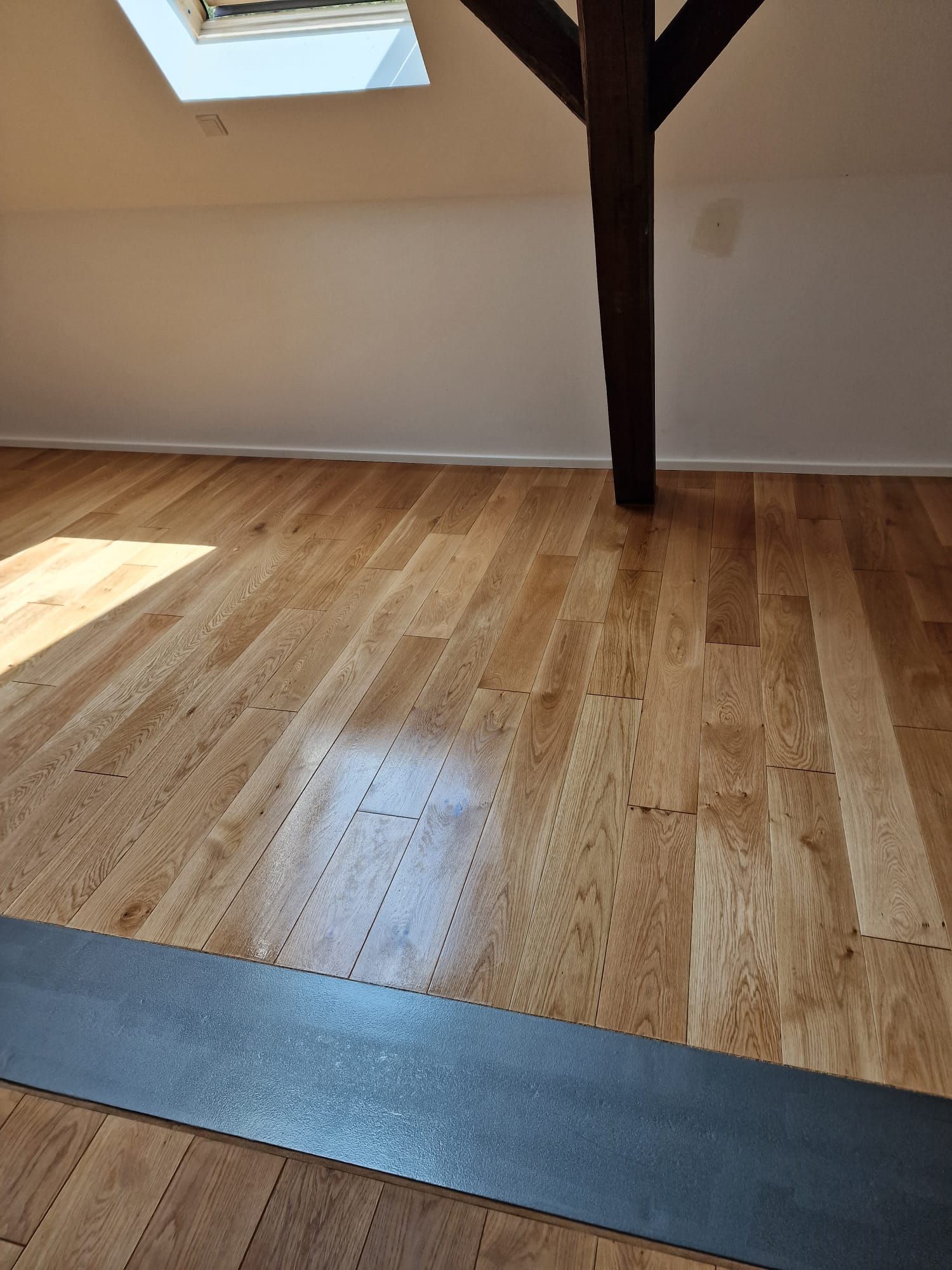 Der Raum verfügt über einen Holzboden mit Stützbalken und Oberlicht. Im Vordergrund ist ein dunkelgrauer Streifen auf dem Boden zu sehen.