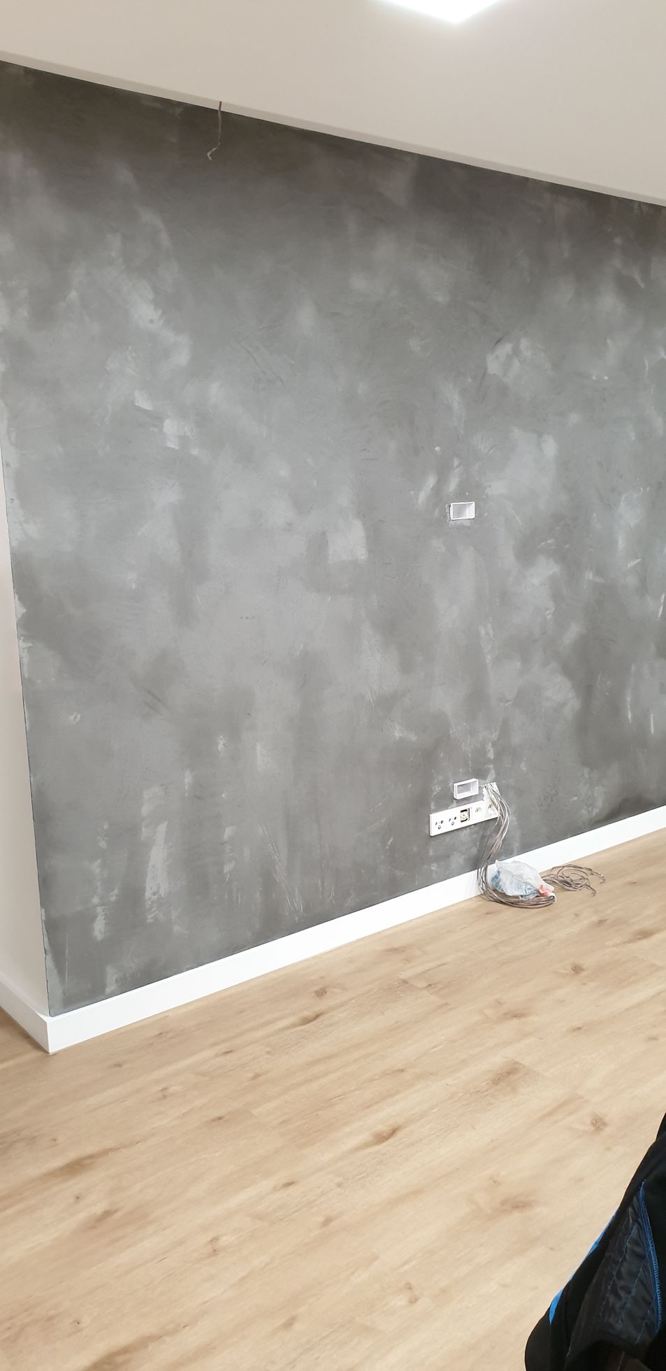 Graue, strukturierte Wand mit weißen Zierleisten und Holzboden. Steckdosen sind sichtbar.