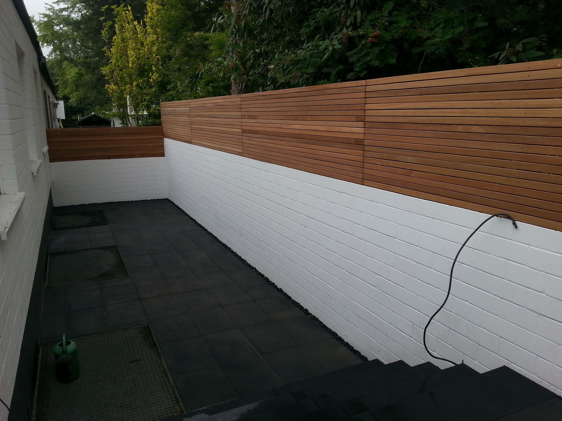 Holzzaun an weißer Backsteinmauer, schwarzer Bodenbelag, grüne Bäume im Hintergrund.