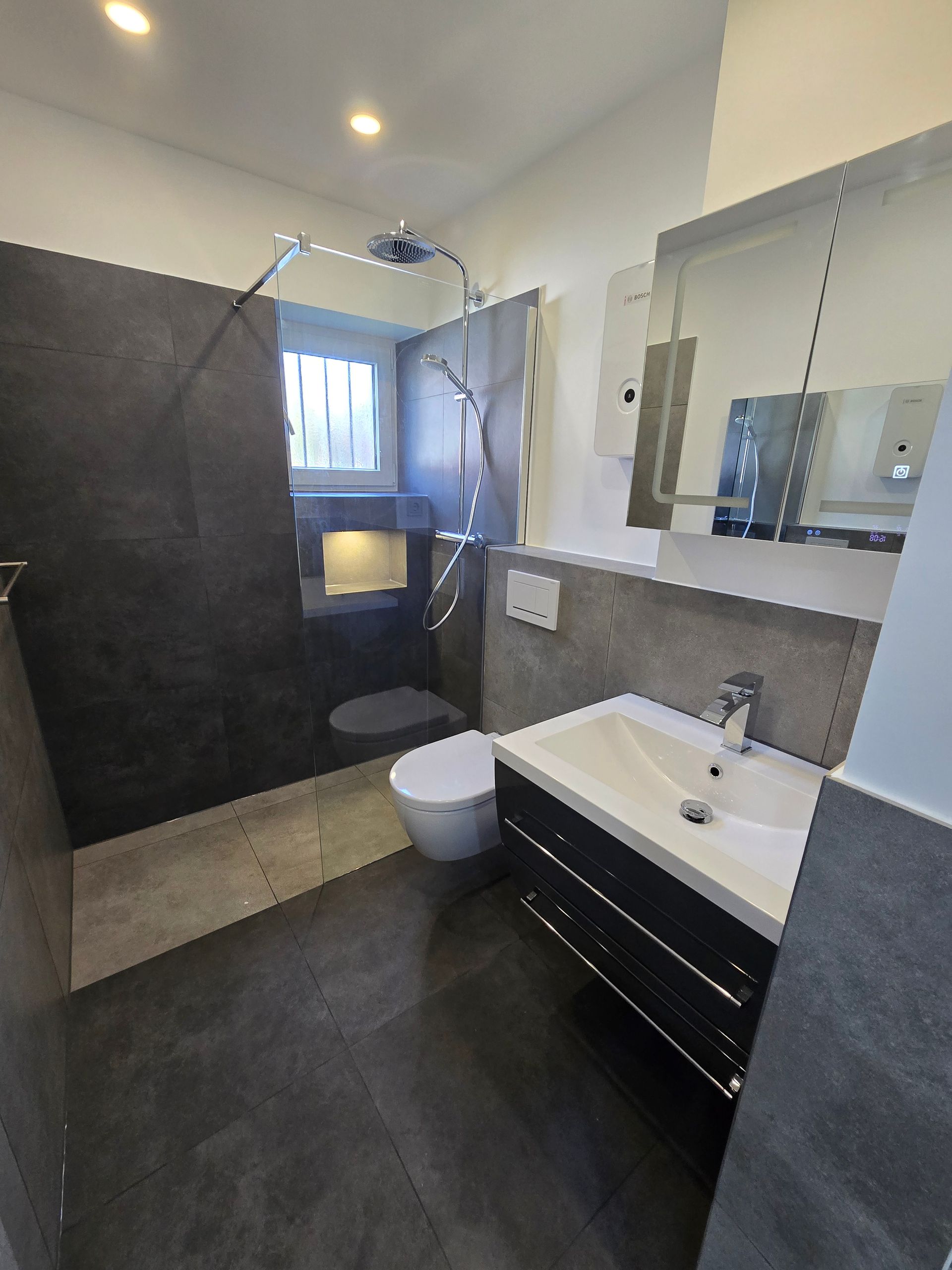 Modernes Badezimmer mit dunkelgrauen Fliesenwänden, Toilette, Waschbecken und Dusche.