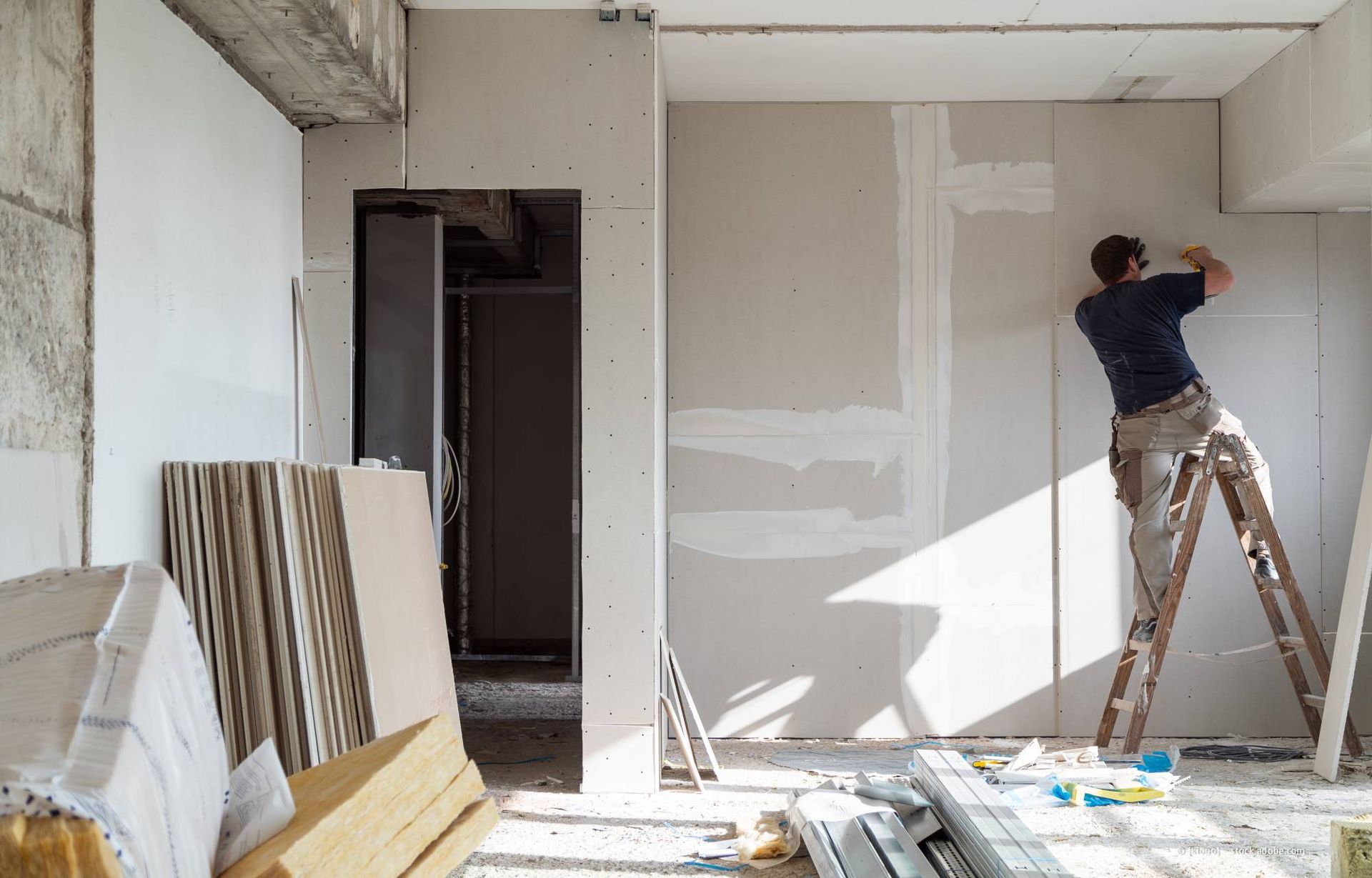 Ein Mann steht auf einer Leiter und montiert Gipskartonplatten in einem im Bau befindlichen Raum.