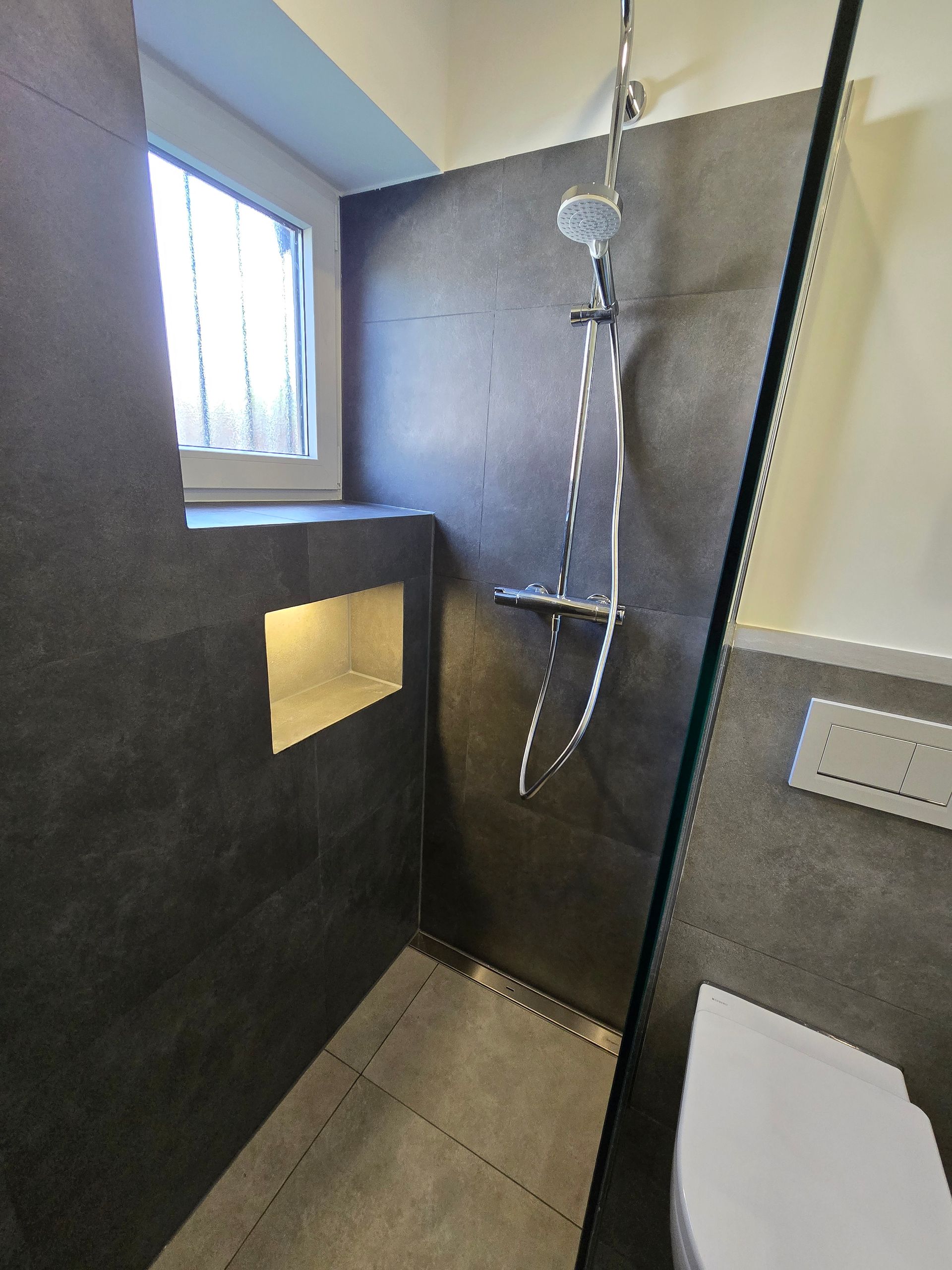 Dunkel geflieste Dusche mit Glaswand, eingebautem Regal, Duschkopf und Fenster.