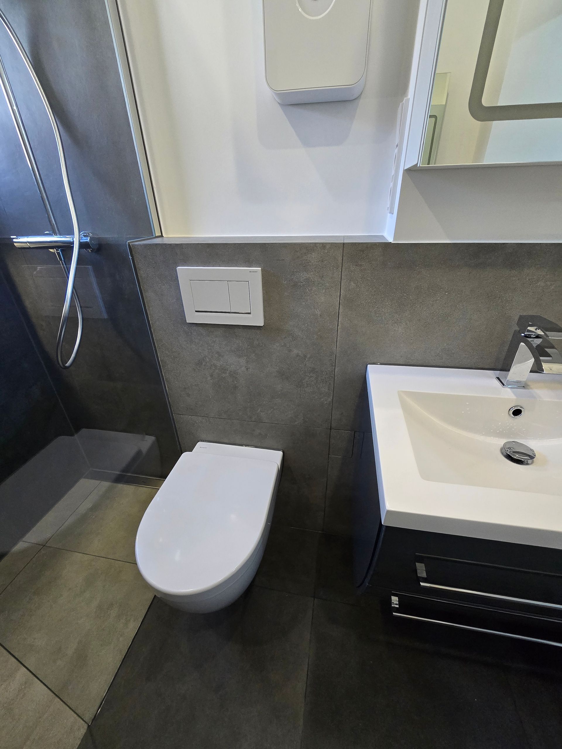 Modernes Badezimmer mit Toilette, Waschbecken und grau gefliester Wand. Weiße Toilette mit Spülknopf.