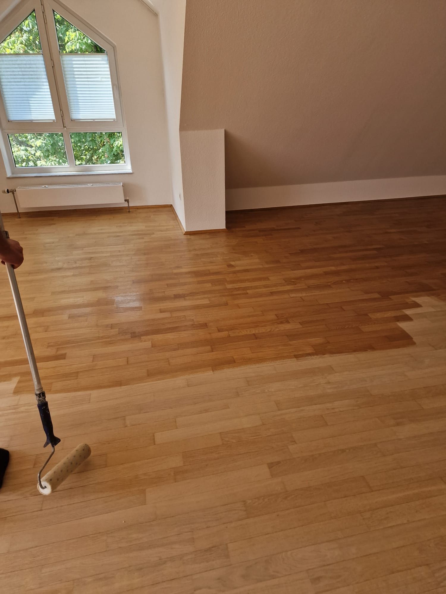 Eine Person trägt Holzbeize auf einen hellen Hartholzboden in einem Raum mit Dachschräge und Fenster auf.