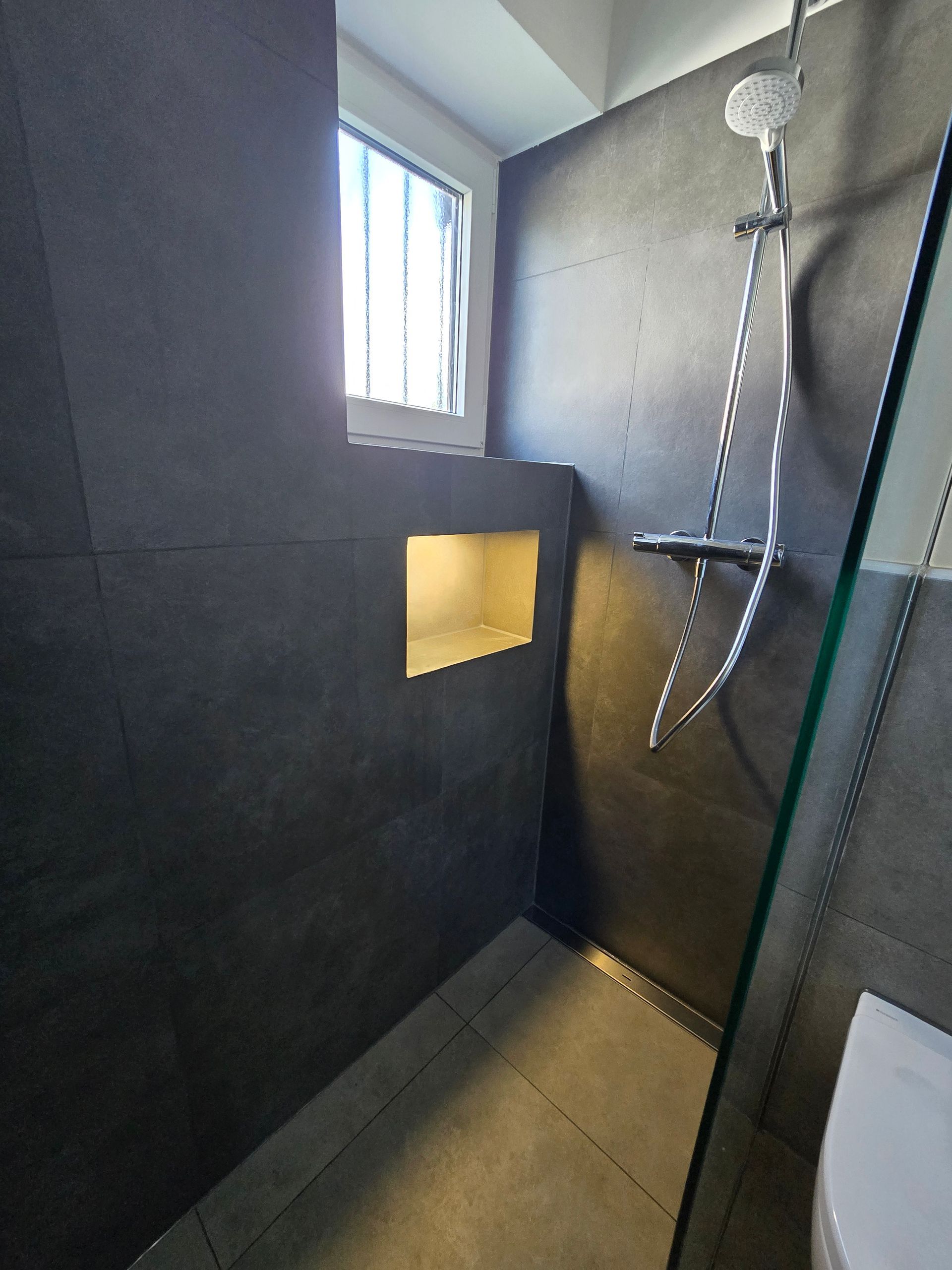 Moderne Dusche mit dunkelgrauen Fliesenwänden, einer eingelassenen Ablage und einer Duschabtrennung aus Glas.