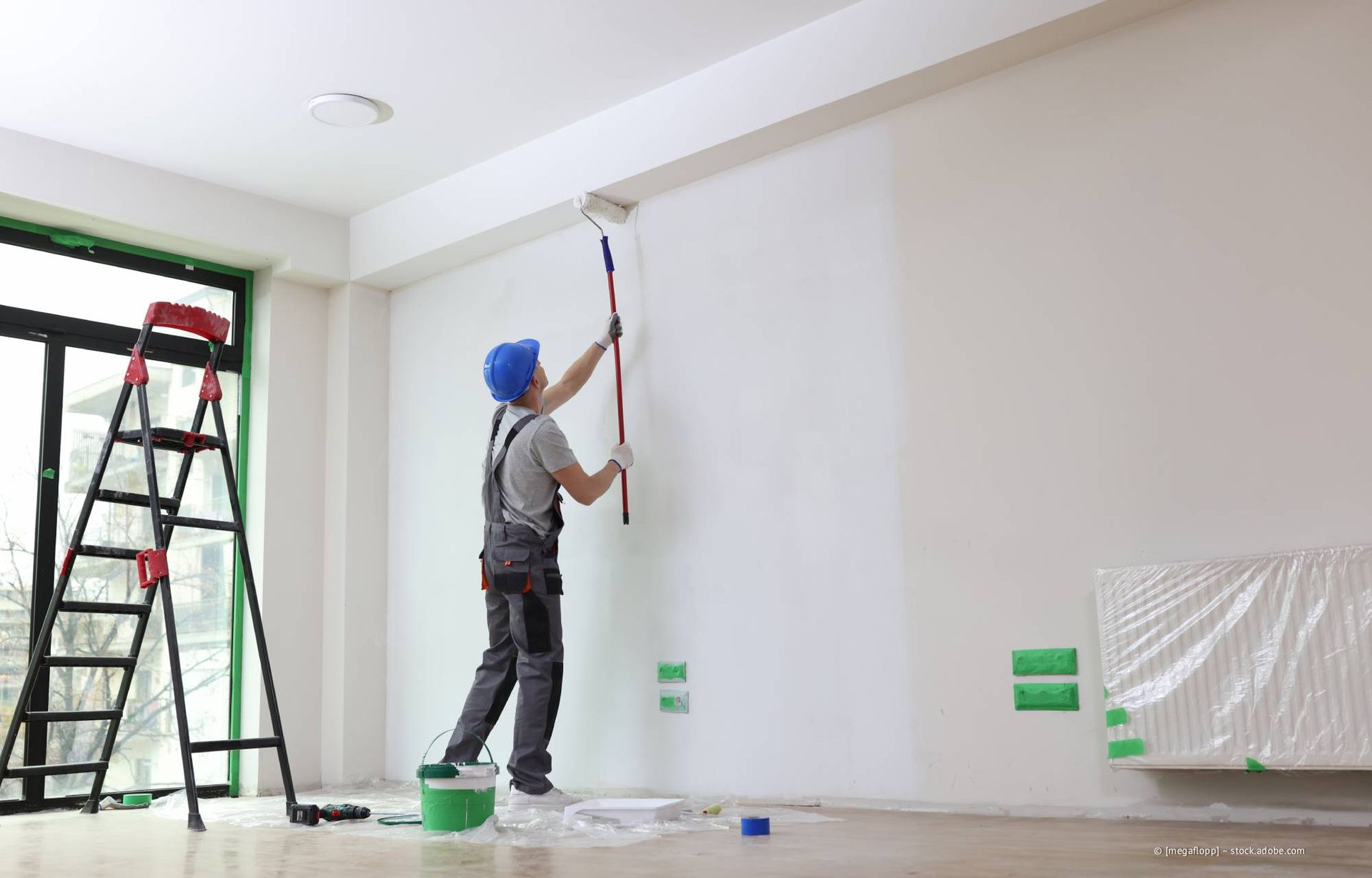 Maler in Overall und Schutzhelm streicht mit einer Farbrolle eine weiße Wand.