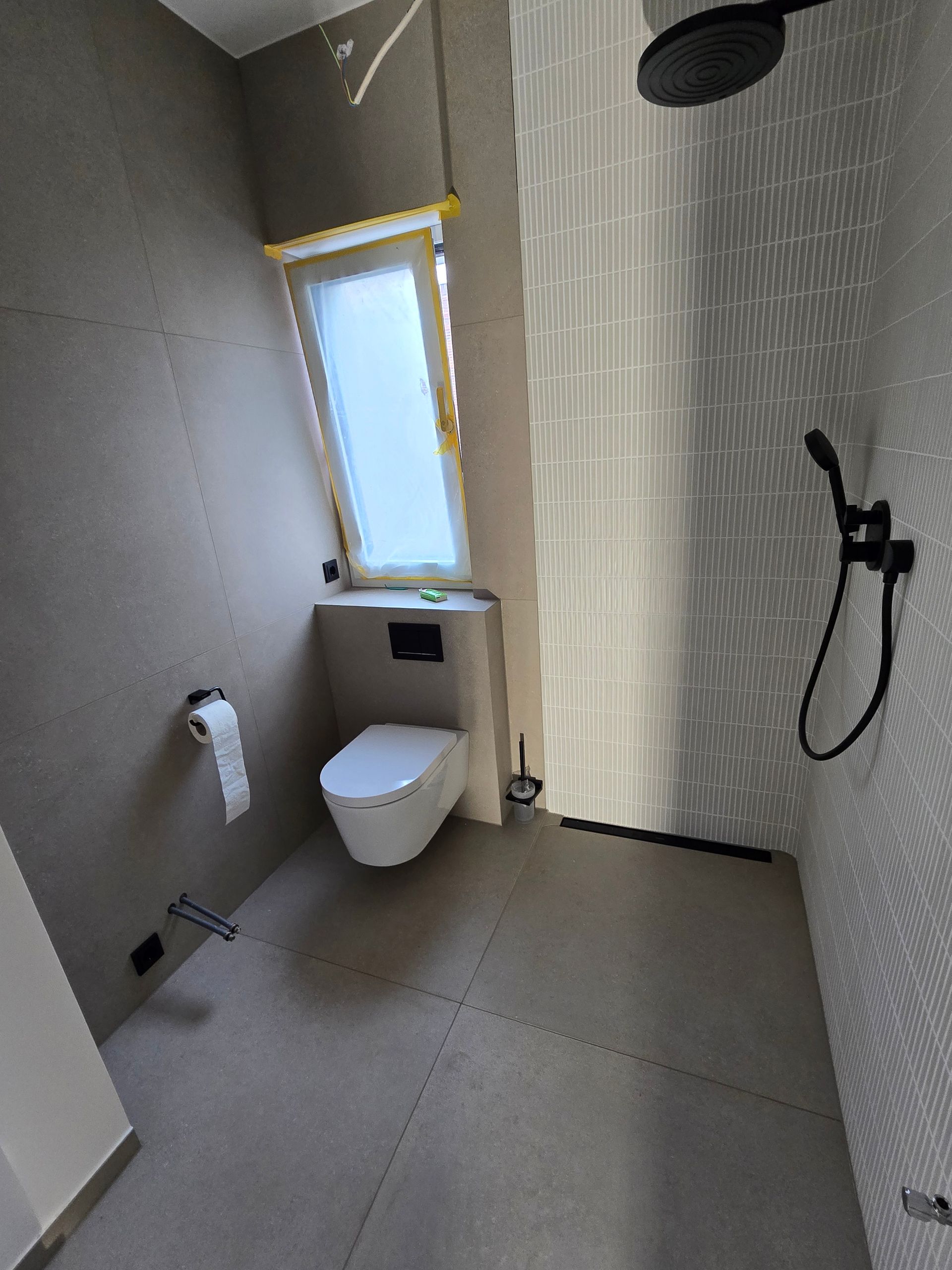 Modernes Badezimmer mit grau gefliesten Wänden und Boden, Toilette unter einem kleinen Fenster, schwarze Duscharmaturen.