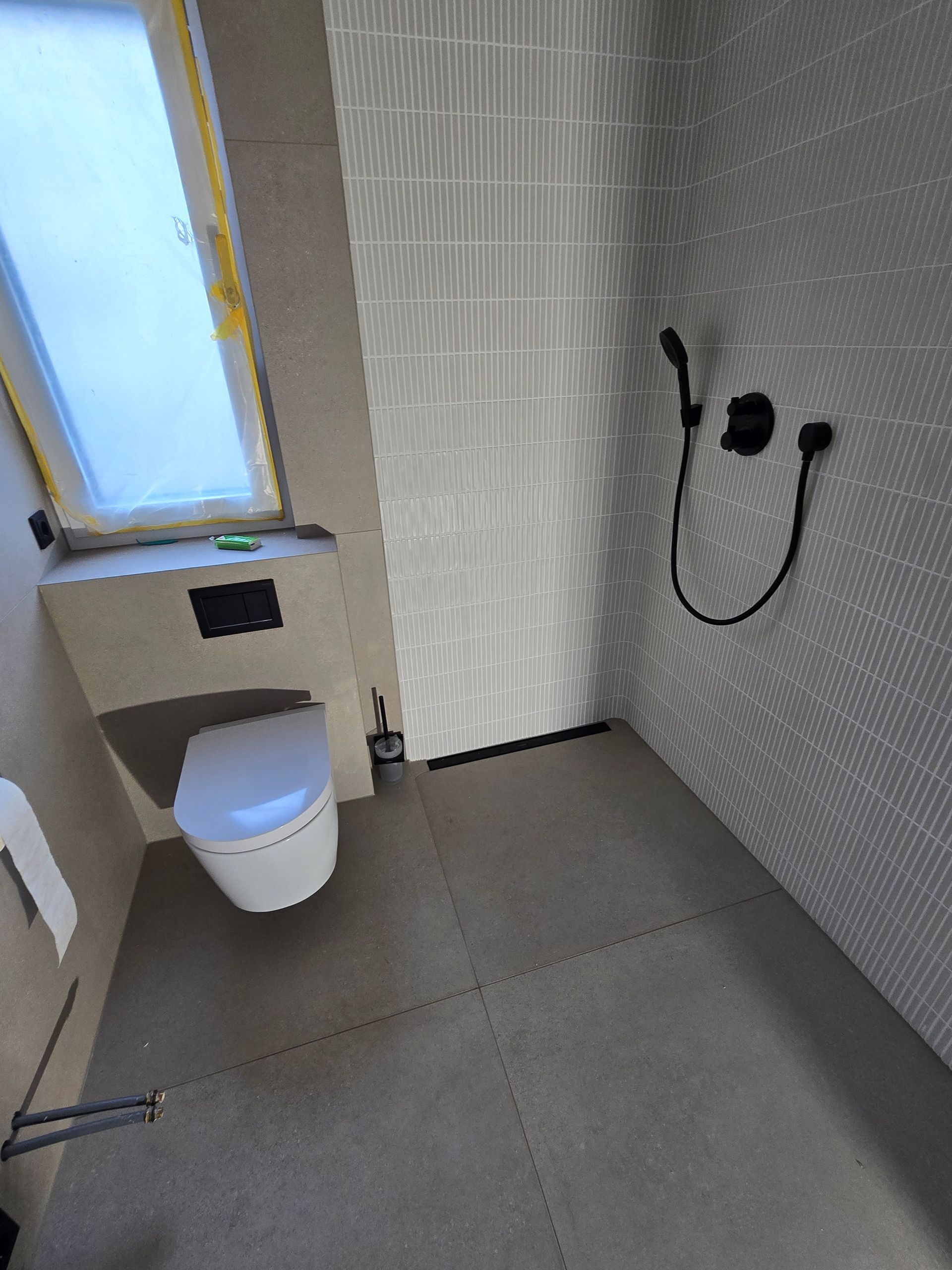 Modernes Badezimmer mit Toilette, Dusche, grauen Fliesen an Wänden und Boden sowie einem Fenster.